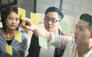 Three people brainstorm with sticky notes on glass wall.