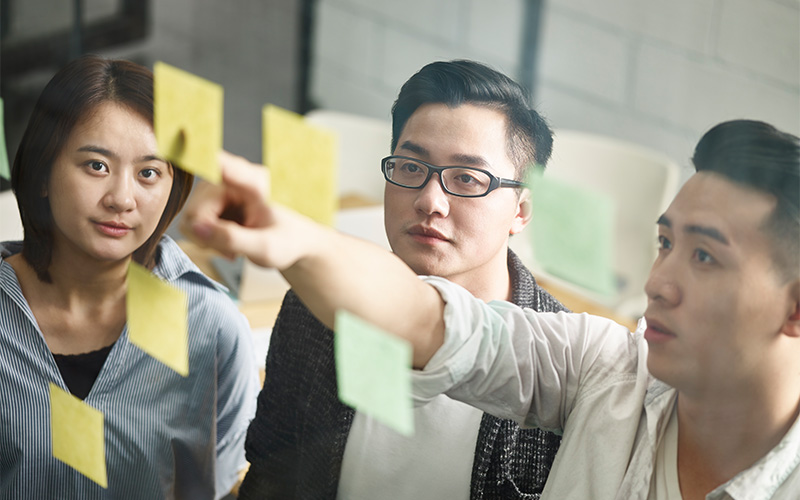 Three people brainstorm with sticky notes on glass wall.